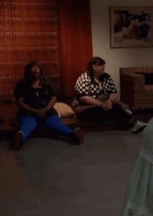 two women sit on a couch in a living room looking up