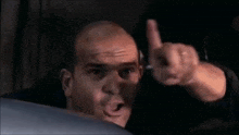a bald man is sitting in a car and pointing his finger at the camera .