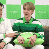 a man in a green and white striped shirt is holding a green pillow with iq.com written on the wall behind him