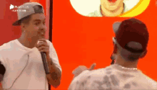 a man in a hat is talking into a microphone while another man looks on