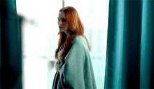 a woman with red hair is wrapped in a blanket and looking out a window .
