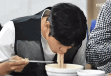 a man is eating noodles with chopsticks from a bowl with a mbc logo behind him