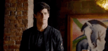 a man in a black jacket is standing in front of a brick wall and a painting of a horse .
