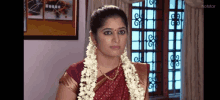 a woman in a red sari is standing in front of a window with a garland of flowers in her hair .