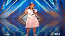a little girl singing into a microphone on a stage with the nbc logo behind her