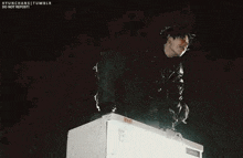a man in a black jacket is standing on top of a white refrigerator .