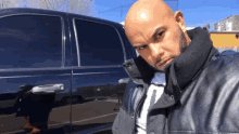 a bald man in a black leather jacket stands in front of a black car