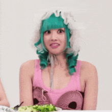 a woman with green hair and a pink tank top is sitting at a table with a plate of salad .