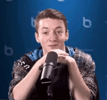 a young man in a plaid shirt is sitting in front of a microphone with his hands folded .