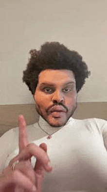 a man with curly hair and a beard is wearing a white turtleneck and making a funny face with his finger .