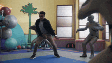 a man in a black jacket is dancing in a gym with a palm tree in the background