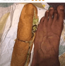 a person 's foot next to a sub sandwich on a napkin