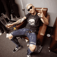 a man in a black shirt with a skull on it sits in a chair giving a thumbs up