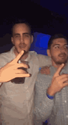 two men are standing next to each other in a dark room and one is holding a bottle of beer .