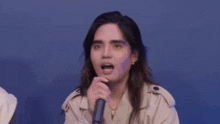 a woman in a trench coat is smiling while sitting in front of a blue background .