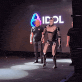 a man in a black shirt stands next to a robot in front of an idol sign