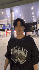 a man wearing a bahz valley t-shirt stands in front of a sign for korea
