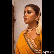 a woman in a yellow saree and gold jewelry is standing in front of a wall .