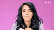 a woman with blue hair stands in front of a pink background that says elite daily on it