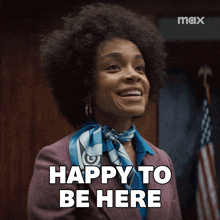 a woman in a suit and scarf is smiling and says " happy to be here "