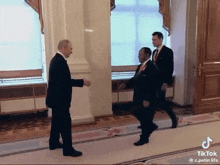 a man in a suit walks down a hallway next to another man in a red tie