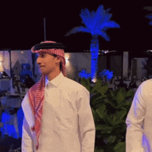 a man wearing a head scarf and a white shirt is standing in front of a palm tree