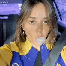 a woman blowing a bubble in a car wearing a yellow and blue jacket