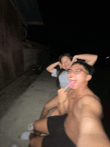 a man without a shirt is sitting next to a woman who is sticking her tongue out at the camera