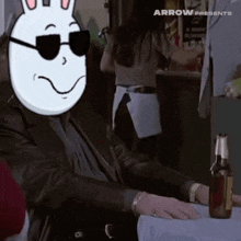 a man wearing sunglasses and a bunny mask sitting at a table with a bottle of beer .
