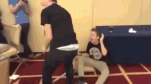 a man in a black shirt is dancing in a room with another man sitting on the floor .