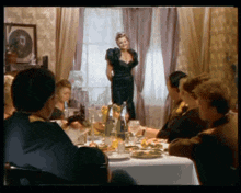 a woman in a black dress is standing in front of a group of people at a table