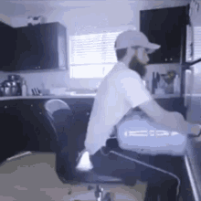 a man with a beard is sitting in a chair in a kitchen using a computer .