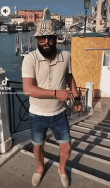 a man with a beard wearing a plaid hat and sunglasses stands in front of a river