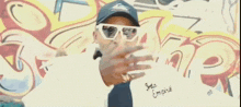 a man wearing sunglasses and a hat stands in front of a wall with graffiti on it .
