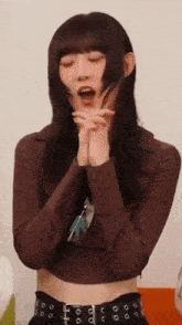 a woman in a brown crop top and black belt is making a funny face with her hands on her face .