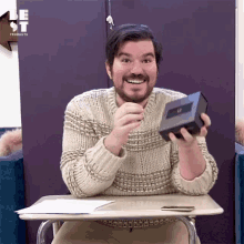 a man in a sweater is sitting at a desk holding a box with the letter t on it