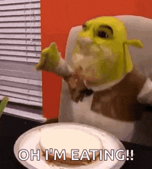 shrek is sitting at a table with a plate of food and eating .