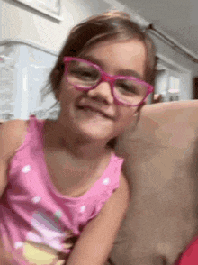 a little girl wearing pink glasses is sitting on a couch and smiling .