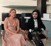 two women are sitting in director 's chairs and one is wearing a pink dress
