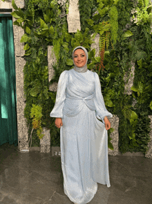 a woman in a light blue dress and hijab stands in front of a wall of greenery