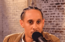 a man in a brown jacket is talking into a microphone in front of a brick wall