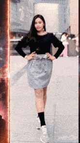 a woman wearing a black shirt and a grey skirt stands on a sidewalk with her hands on her hips