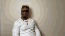 a man wearing sunglasses and a white sweater is sitting against a wall