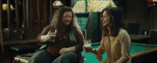 two women are sitting at a pool table and one of them is holding a glass of beer