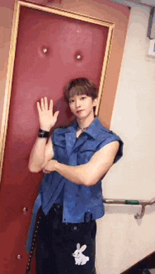 a man in a blue vest is standing in front of a red door and waving his hand .