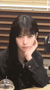 a young girl with long black hair is sitting at a table with her hand on her chin .