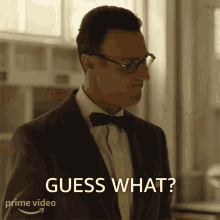 a man wearing glasses and a bow tie says " guess what "