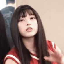 a girl with long black hair and bangs is wearing a red shirt and waving .