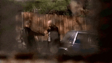 two men are standing next to each other in front of a car and smoke is coming out of the car .
