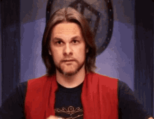 a man with long hair and a beard is wearing a red vest and a black t-shirt .